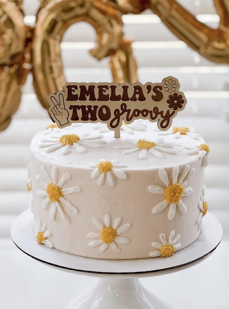 a white cake with daisies on it and a wooden sign that says,'emelia's two grooy '