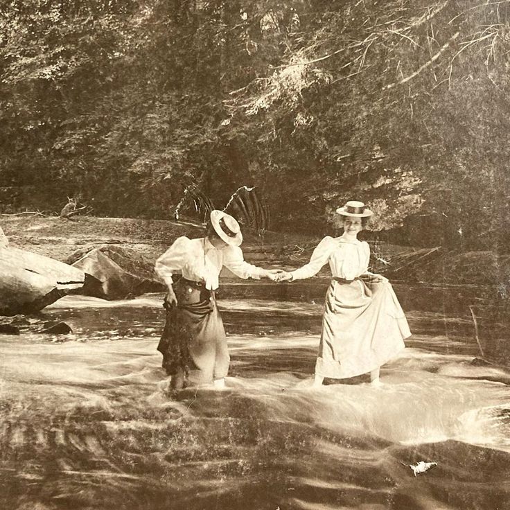 two women in dresses and hats crossing a river with their arms around each other as they walk through the water