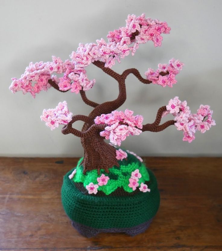 a crocheted bonsai tree with pink flowers in a green pot on a wooden table