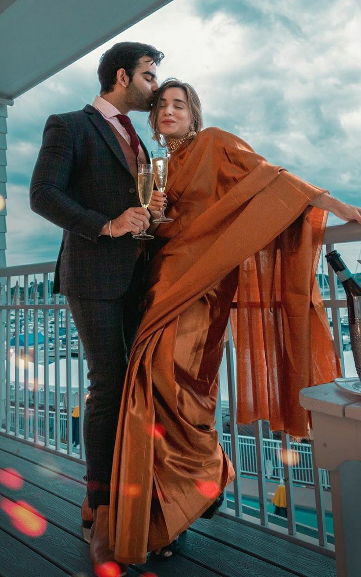 a man and woman standing on a deck with champagne glasses in their hands, holding each other's arms