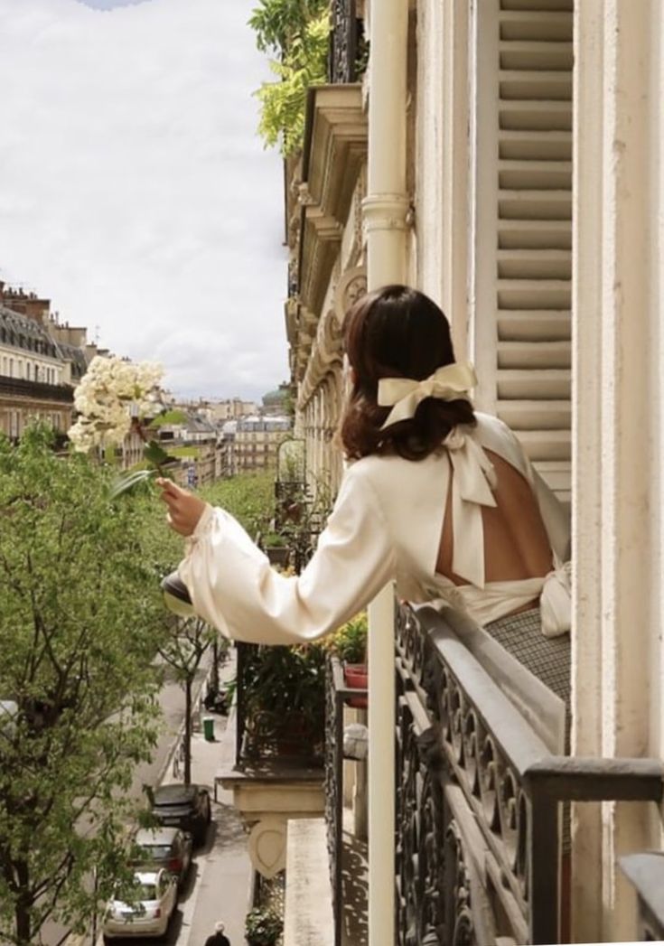 a woman standing on a balcony holding a bouquet of flowers in her hand and looking out the window