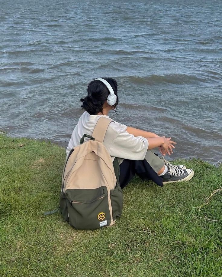 a woman sitting on the grass by the water with her back to the camera, looking at the water