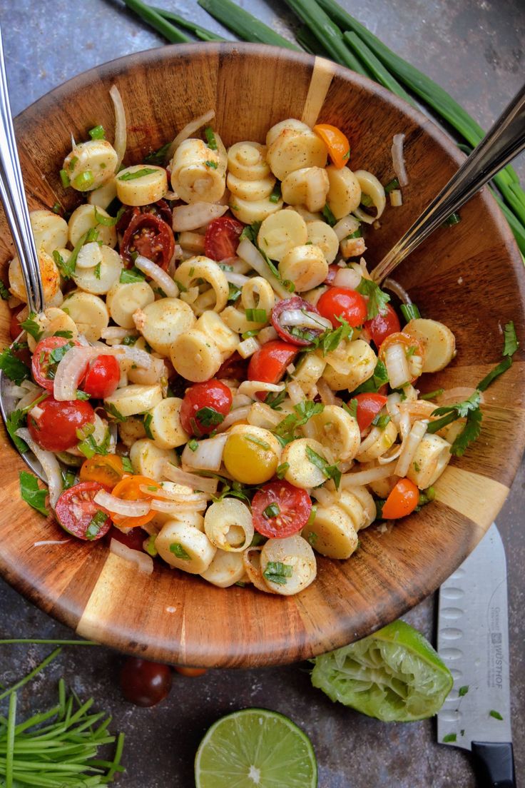 a wooden bowl filled with pasta salad next to green onions and lime wedges on the side