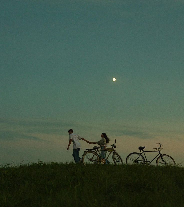 two people holding hands and walking with their bikes in the grass at dusk, while the sun is setting behind them