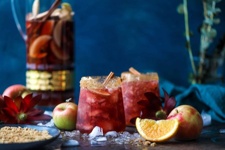 two glasses filled with drinks next to sliced apples and oranges on a table top