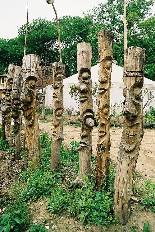 many carved wooden poles in the grass near a dirt road and trees with faces on them