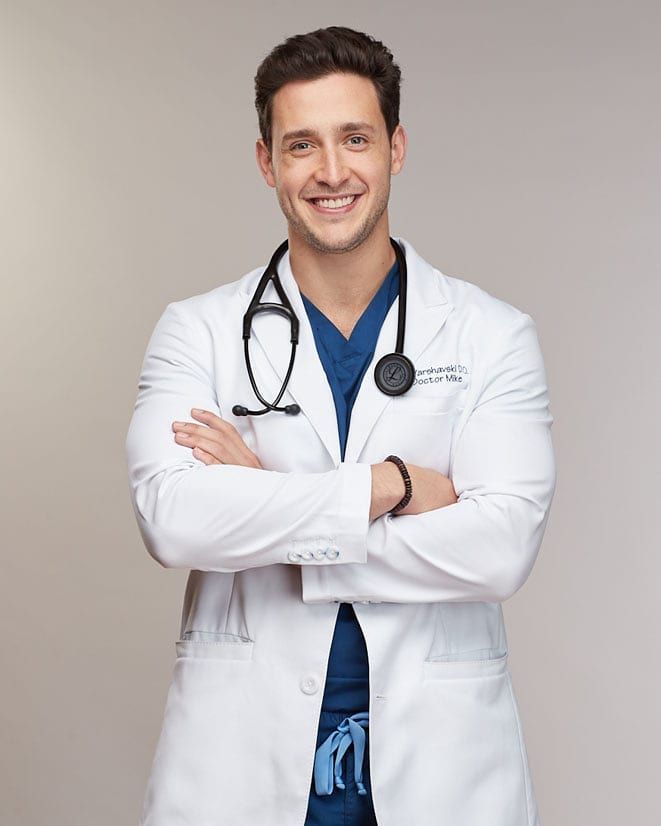 a male doctor in a white coat and blue scrubs is posing for the camera