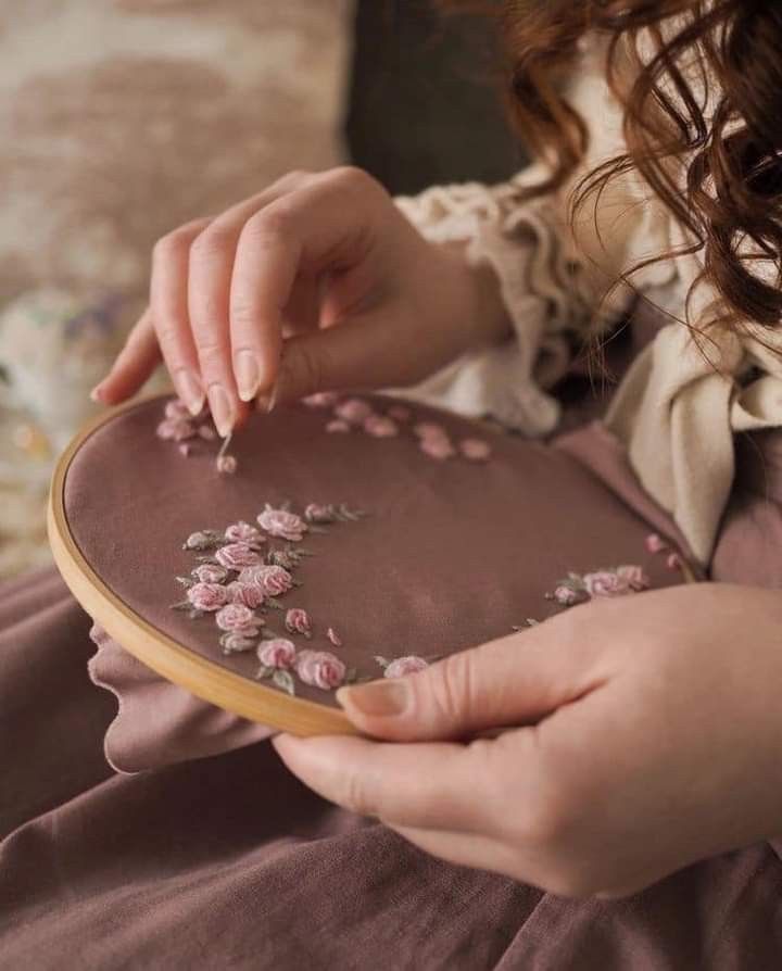 a woman is stitching pink flowers on a brown cloth with a wooden embroidery hoop