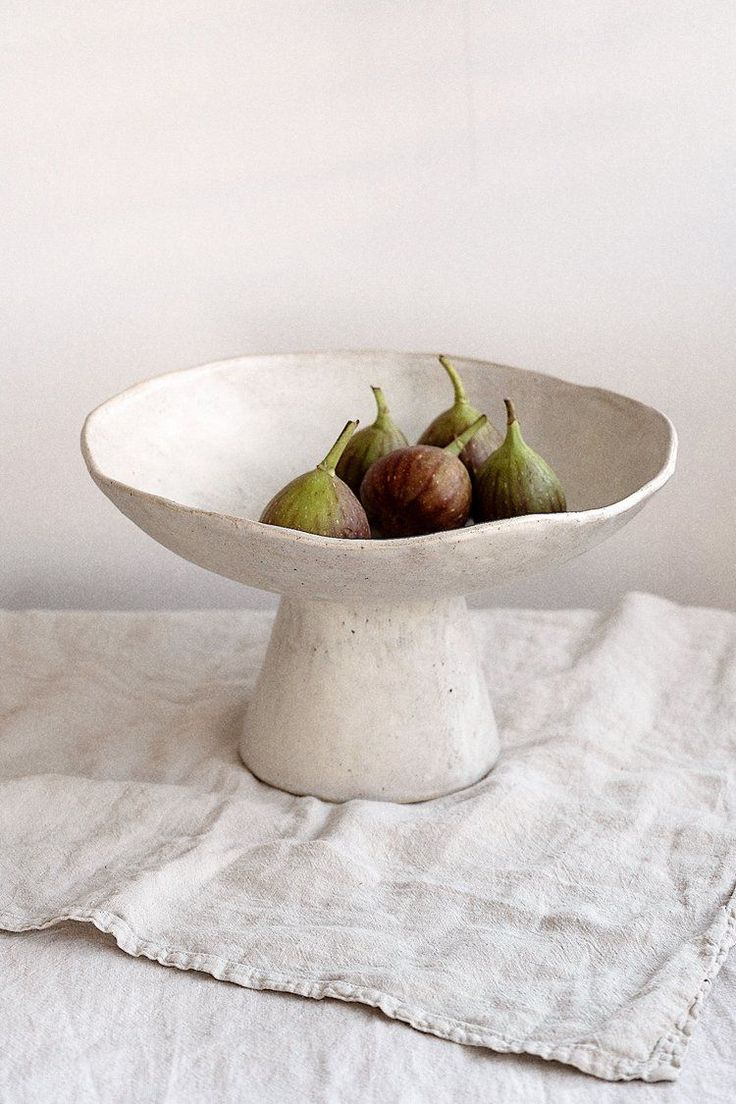 a white bowl filled with figs on top of a cloth covered table next to a wall