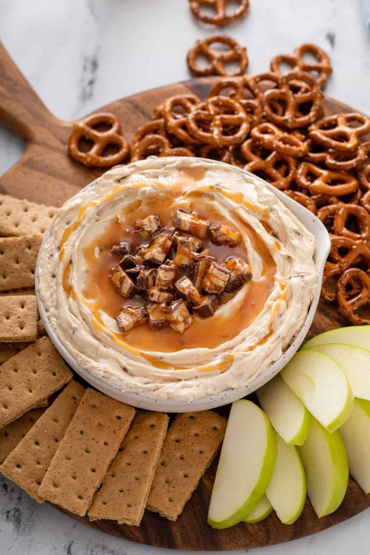 an apple and pretzel dip surrounded by crackers on a wooden platter