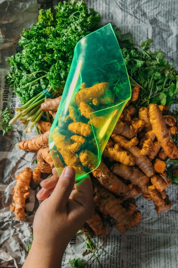 a person holding up a plastic bag filled with carrots and broccoli next to other vegetables