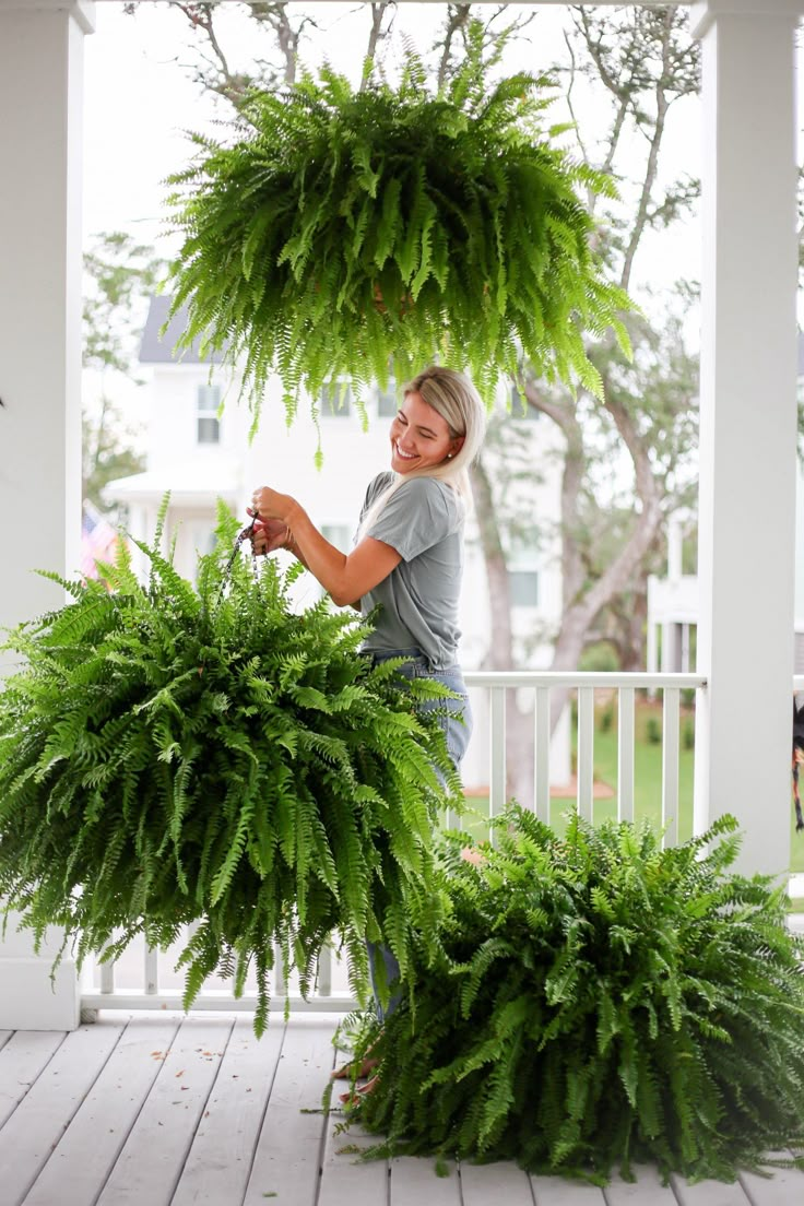 a woman standing on a porch next to two large green plants hanging from the ceiling