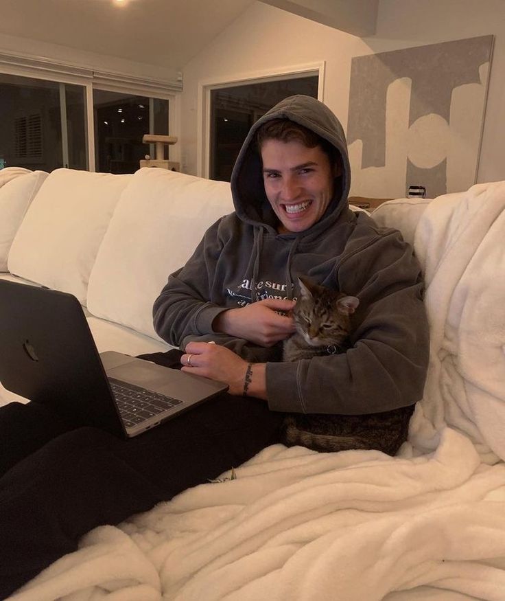 a man sitting on a couch holding a cat and using a laptop computer while smiling at the camera