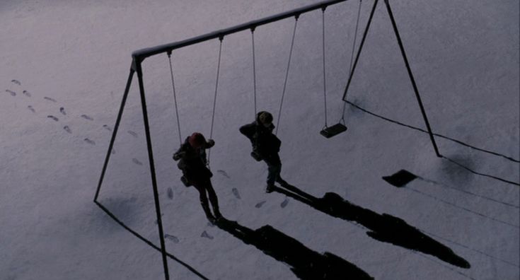 two people on swings in the snow at night, with one person standing next to them