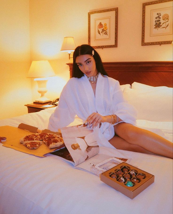 a woman in white robe sitting on bed with pizzas and boxes next to her