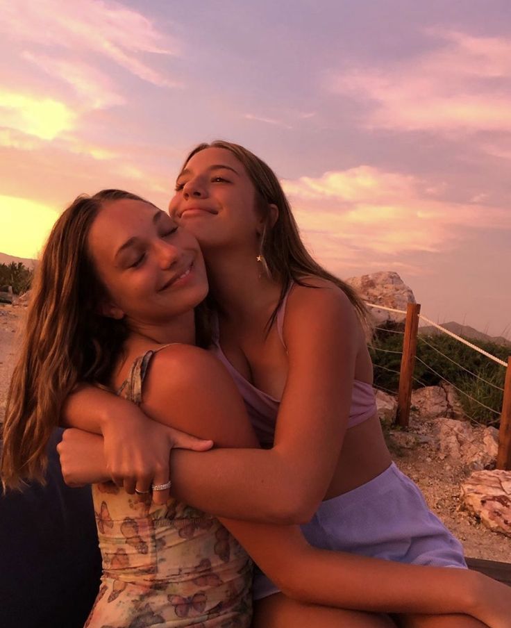 two young women hugging each other on the beach at sunset, with one woman sitting down and another standing up