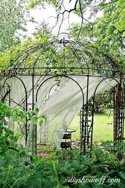 an outdoor gazebo in the middle of some bushes and trees with a white cloth draped over it