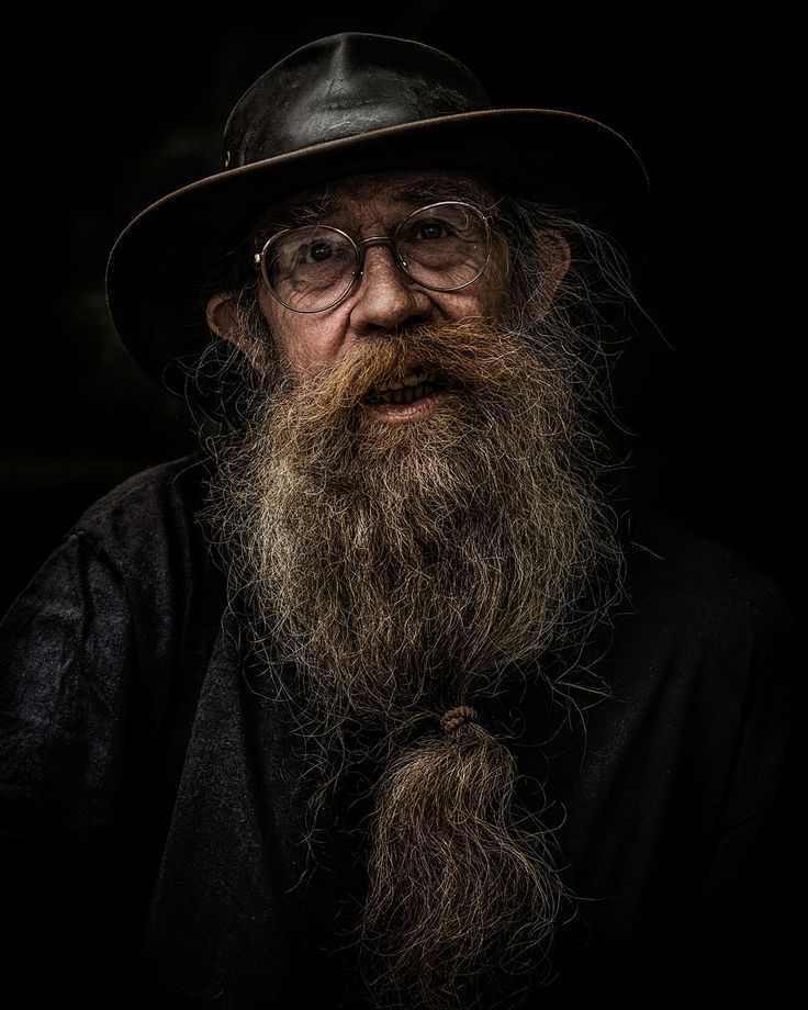 an old man with a long beard wearing a hat and black shirt is looking at the camera