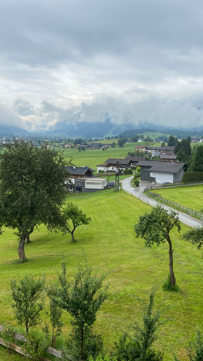 an open field with trees and buildings in the distance on a cloudy day, surrounded by mountains