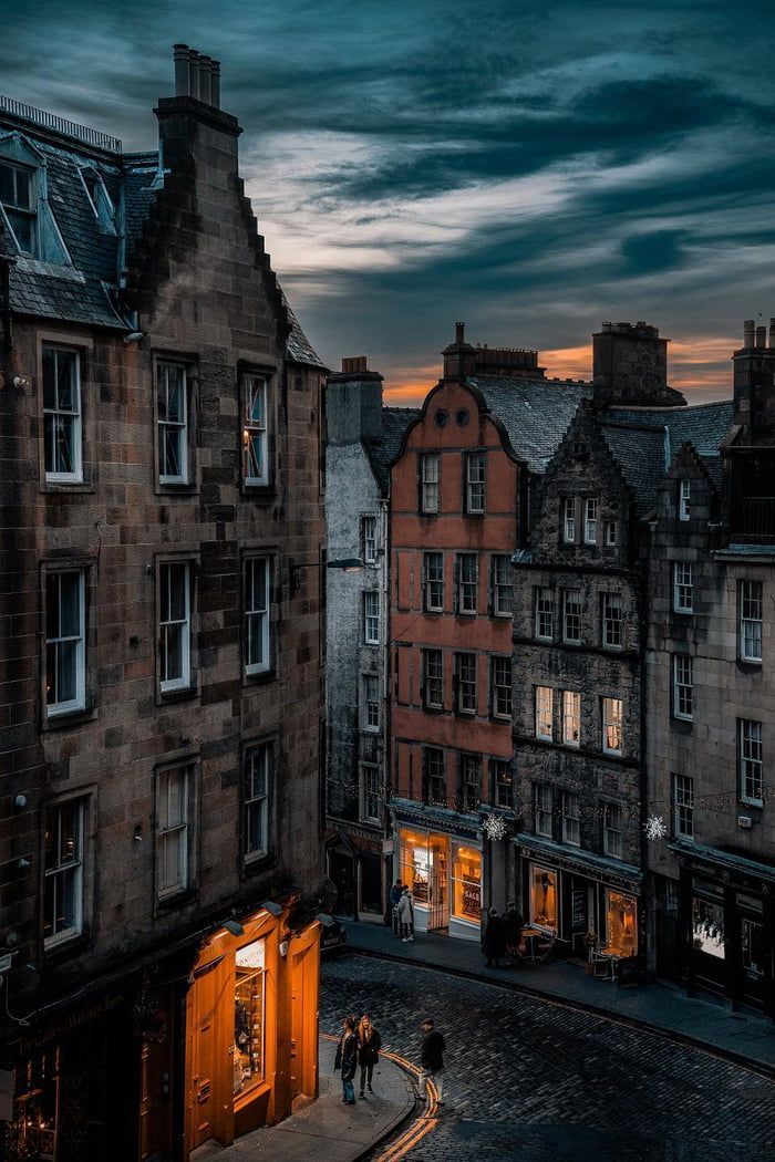 two people are walking down the street in front of some buildings at night with lights on