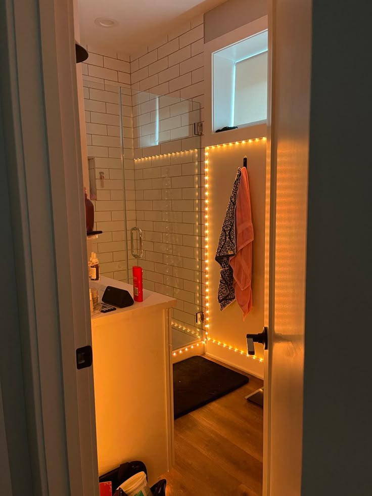 White polished bathroom with glass shower, with soft orange LED lights decorating the edges Bathroom Mirrors Small Space, Cute Bathroom Lighting, Aesthetic Bathroom Shower Ideas, Bathroom Couple Ideas, Bedroom Decor College Apartment Inspiration, Bathroom Small Aesthetic, Aesthetic Bathroom Lights, Mood Lighting For Bathroom, College Apartment Halloween Decor