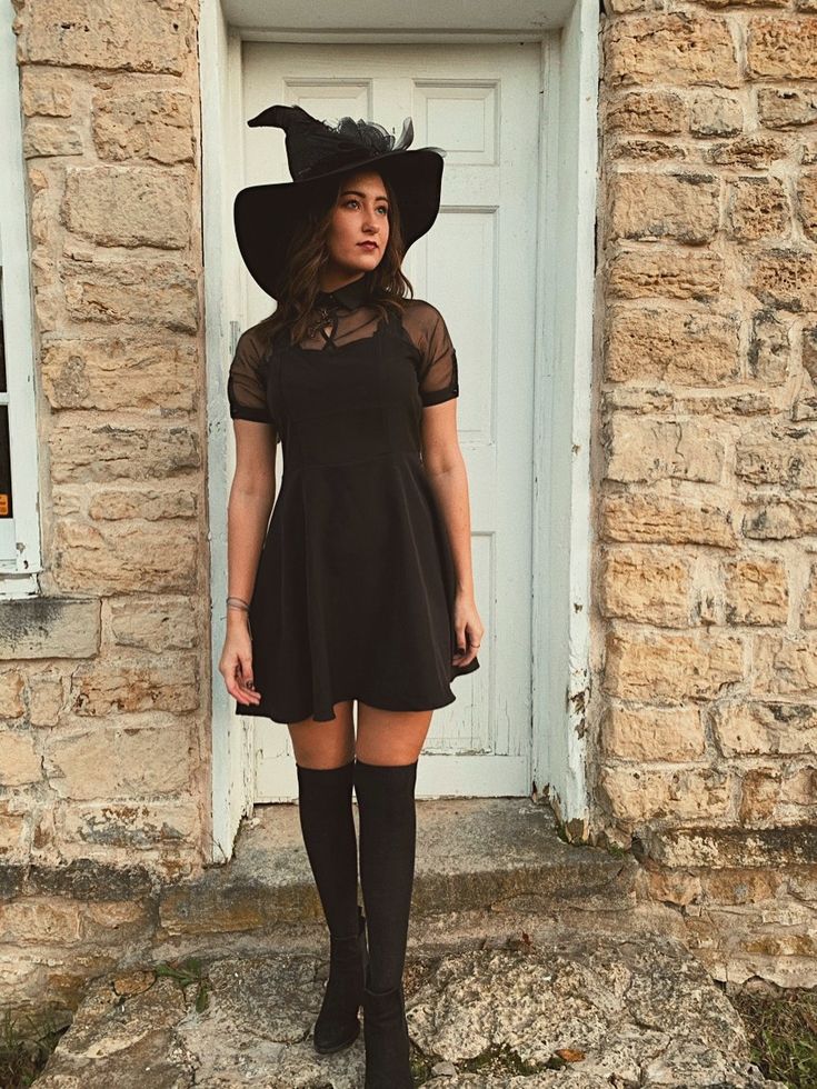 a woman wearing a black dress and hat standing in front of a door with her hands on her hips