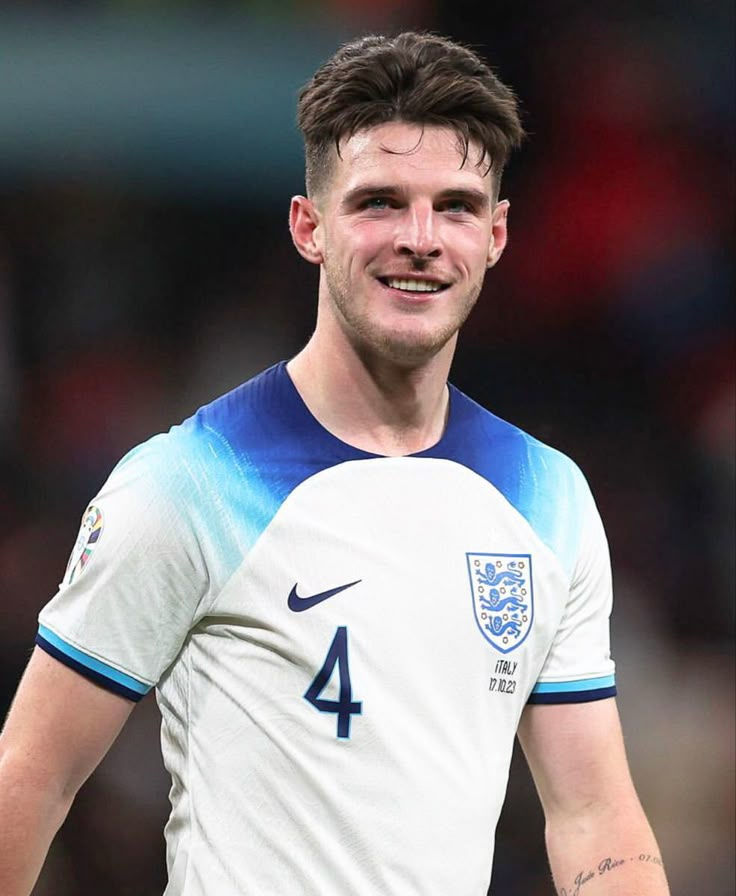 a young man in a white and blue shirt smiles at the camera while standing on a soccer field