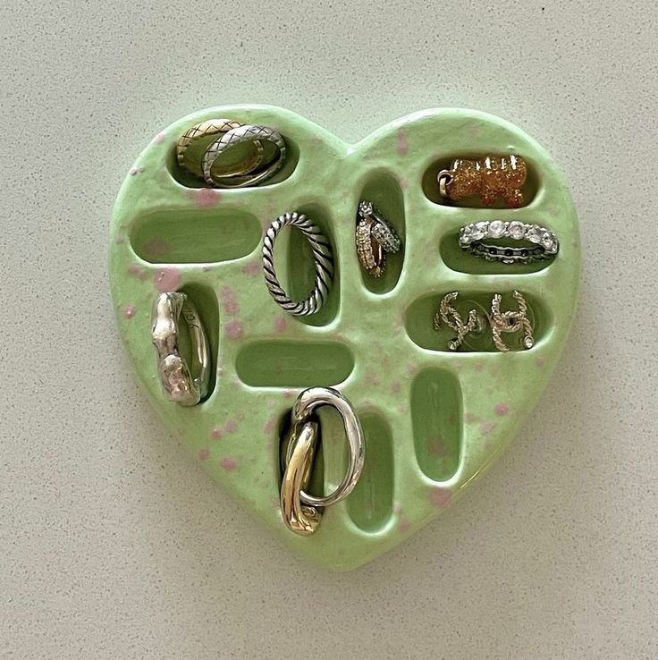 a green heart shaped tray with various rings and jewelry in it on a white surface