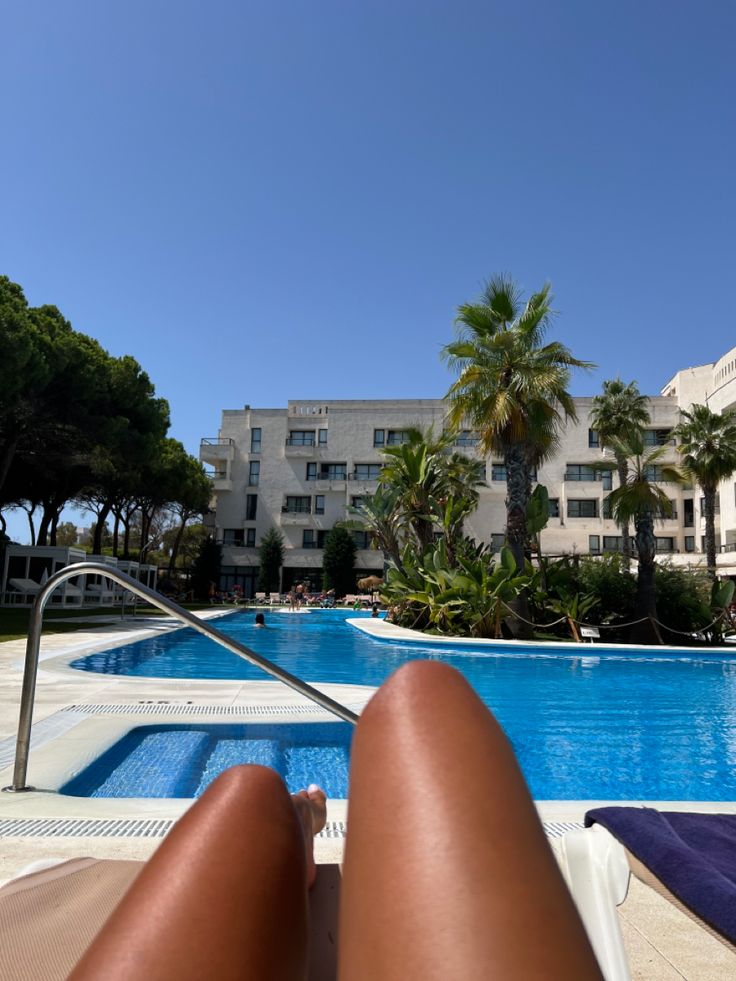 a person laying in a lounge chair next to a swimming pool with palm trees and buildings in the background