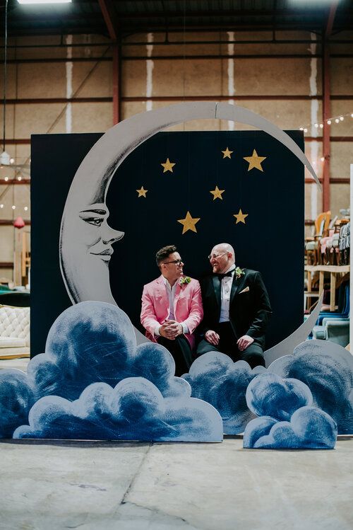 two men sitting on clouds in front of a large moon and stars sign with the sky painted onto it