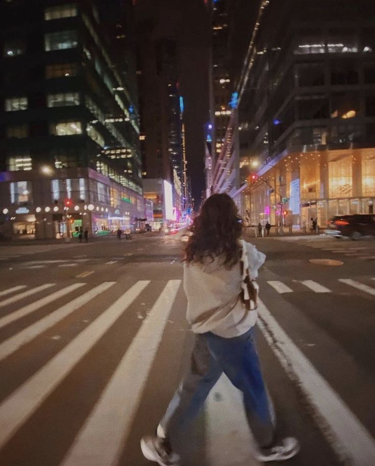 a woman walking across a cross walk in the middle of a city street at night