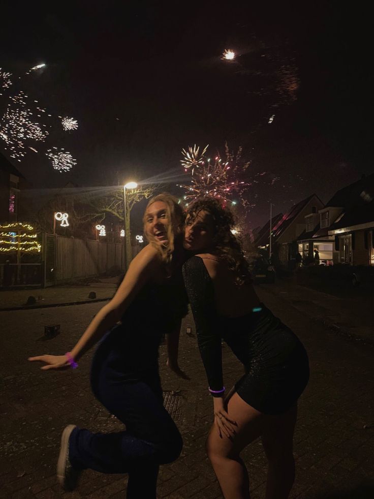 two women are dancing in the street at night with fireworks behind them and onlookers