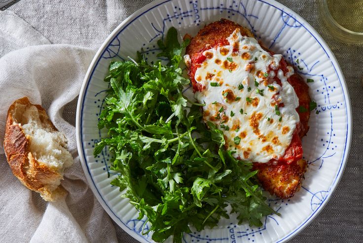 a white plate topped with meat covered in sauce and greens next to a piece of bread