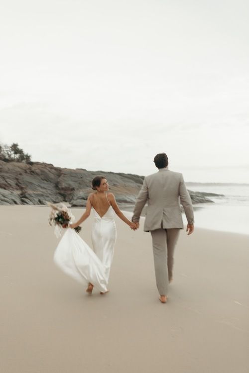 a bride and groom walking on the beach holding hands