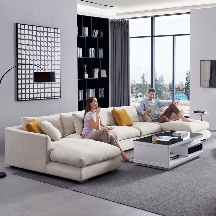 a man and woman sitting on a couch in a living room with large windows looking out onto the water