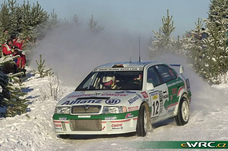 a rally car driving through the snow in front of some evergreens with people watching