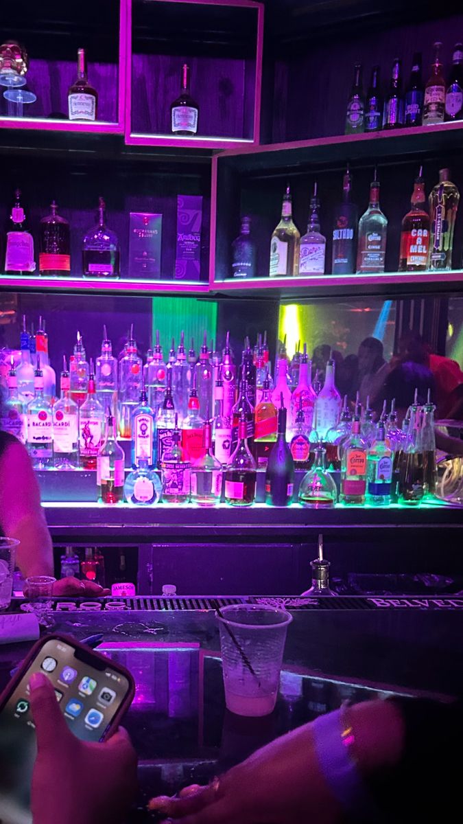 a man sitting at a bar with a cell phone in his hand and bottles on the shelves behind him