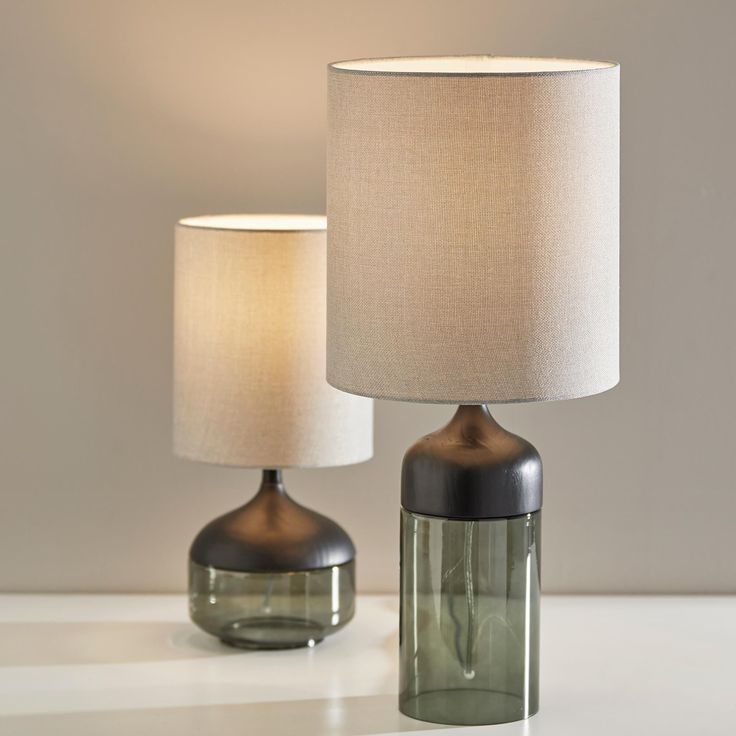 two glass lamps sitting next to each other on a white table with a beige lamp shade