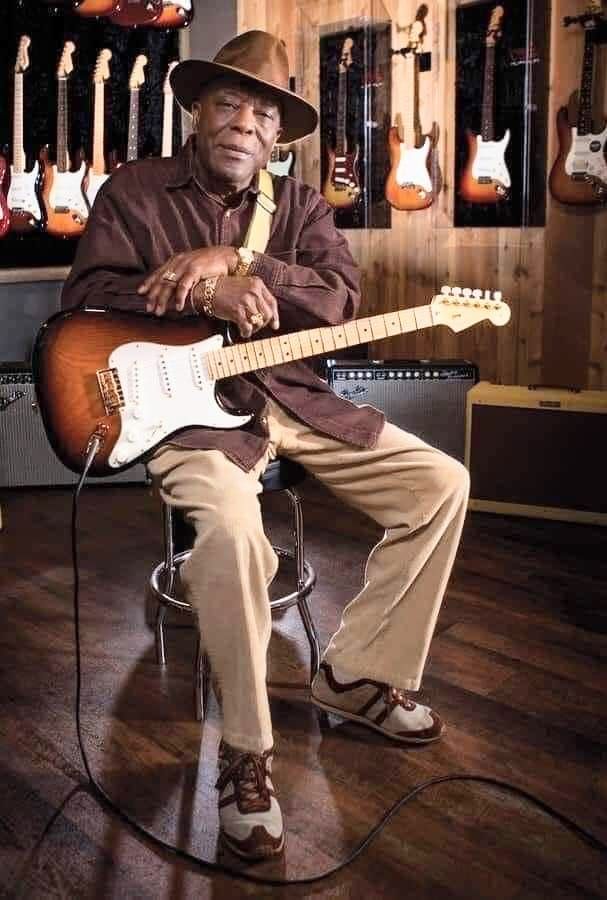 a man sitting in a chair with an electric guitar on his lap and wearing a hat