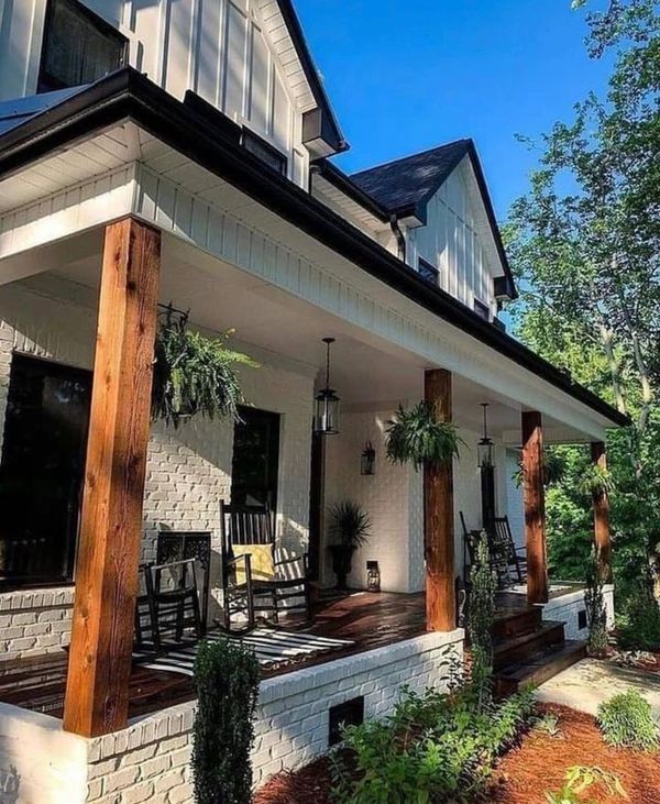 a white house with wooden porches and chairs on the front porch, surrounded by greenery