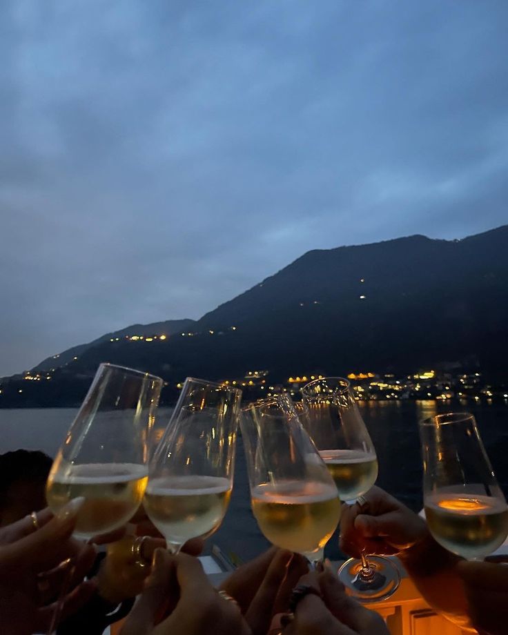 several people toasting with wine glasses in front of the water at night, near a mountain