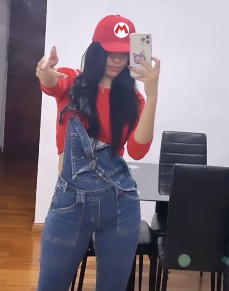 a woman wearing overalls and a red hat taking a selfie with her cell phone