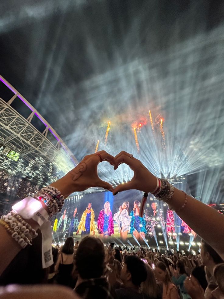 two hands making a heart shape in front of an audience at a concert with fireworks