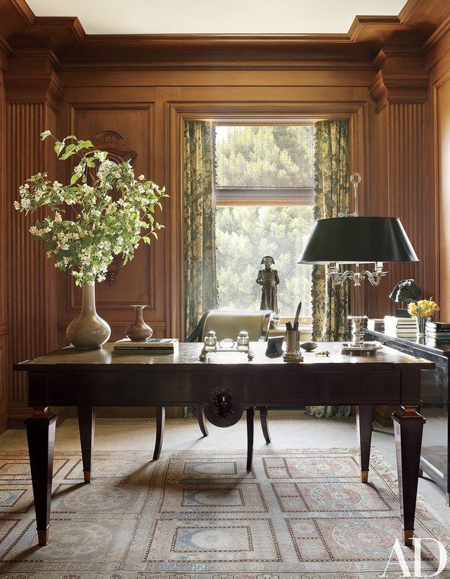 a desk with a lamp, vase and flowers on it in front of a window