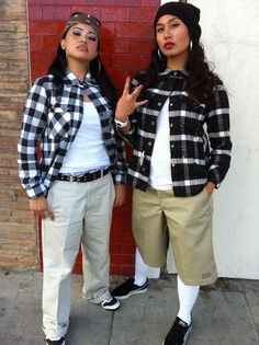 two people standing next to each other in front of a brick wall with one pointing at the camera