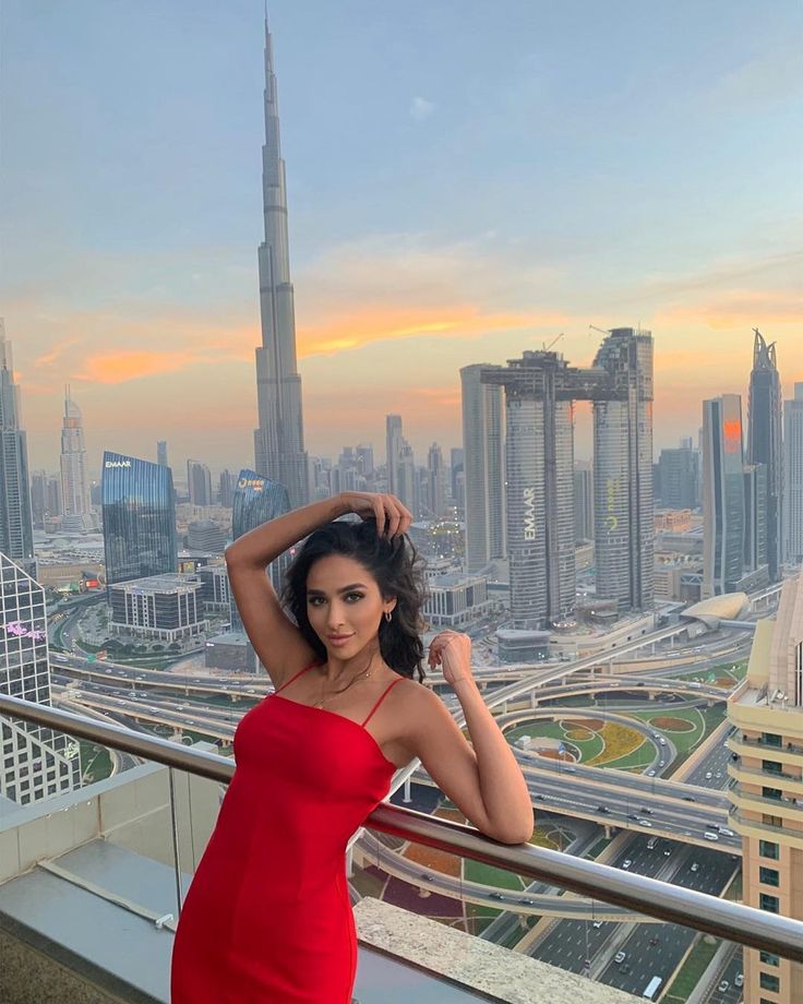 a beautiful woman in a red dress standing on top of a building looking at the camera