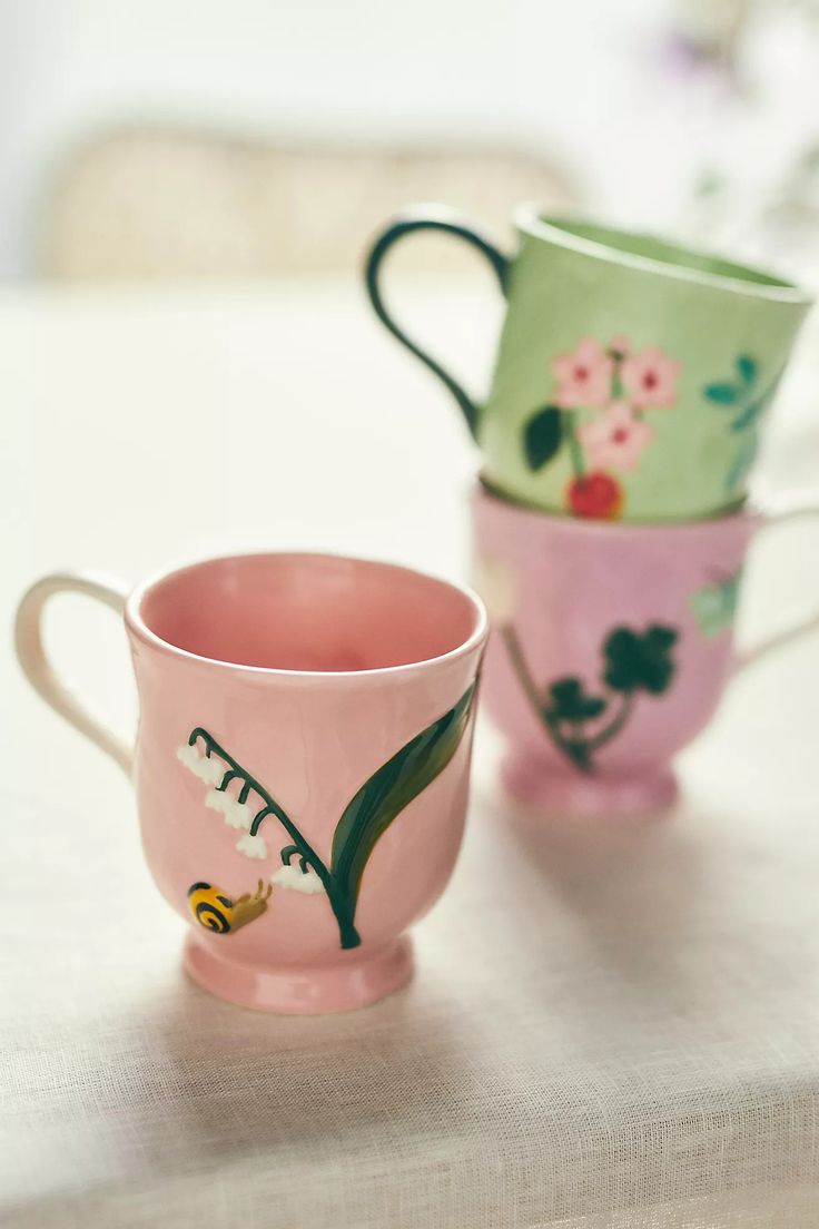 two pink and green mugs sitting on top of a white cloth covered tablecloth