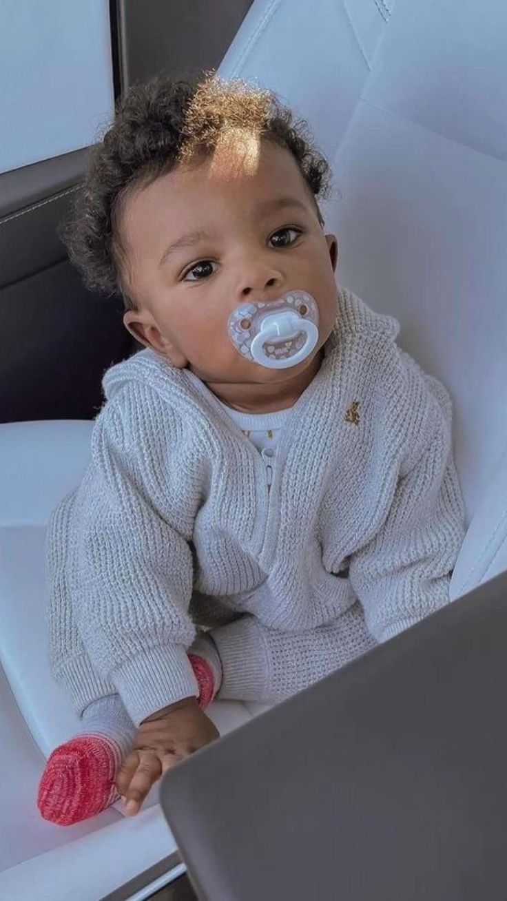 a baby sitting in a white chair with a pacifier on its mouth and looking at the camera