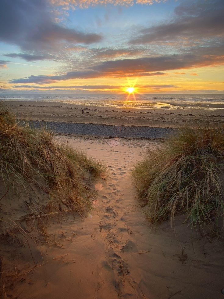 the sun is setting at the beach with footprints in the sand and grass on either side