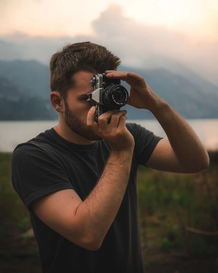 a man holding a camera up to his face in front of a lake and mountains